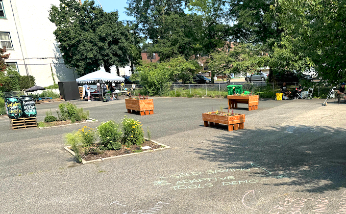 Paved lot with a tent, different raised beds, and flowers blooming in ground beds.