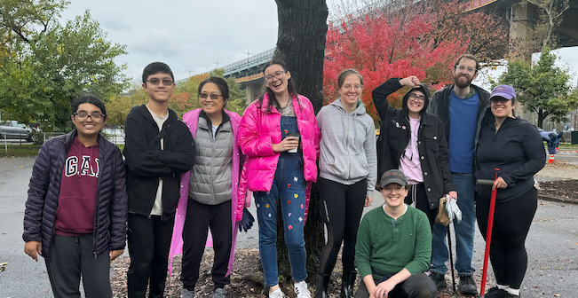 Diverse group of gardeners on a cloudy day, standing in paved lot, smiling in front of autumn trees, holding gardneing tools and assistive devices.